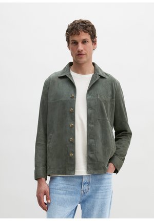 Mann trägt eine dunkelgrüne Wildlederjacke mit Knöpfen, ein weißes T-Shirt mit Rundhalsausschnitt und hellblaue Jeans vor einfachem Hintergrund.