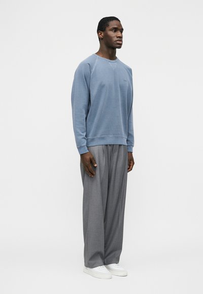 Homme debout portant un sweat-shirt à manches longues bleu clair, un pantalon gris ample et des baskets blanches devant un fond uni.