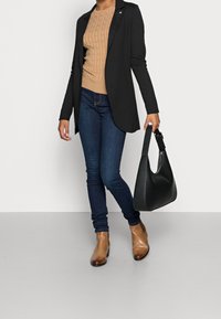 Czarna marynarka na jasnobrązowym sweterku, niebieskie skinny jeans, brązowe botki na kostkę i czarna torebka na ramię. Prosty design i neutralne kolory.