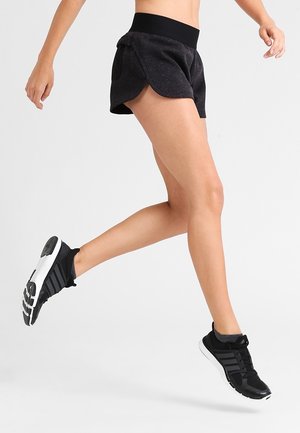 Frau trägt schwarze Sportshorts und schwarze Laufschuhe, eingefangen in der Luft bei einer Sprung- oder Laufbewegung vor neutralem Hintergrund.