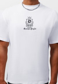 Hvit bomull T-skjorte med en svart logo med krone, utsmykkede kanter, og teksten "Gothic Style" over "EST. 1990." Korte ermer.