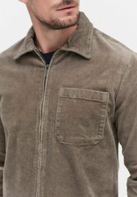 Brown corduroy jacket with a zip front, pointed collar, and single chest pocket. Textured fabric with vertical ribbing pattern.