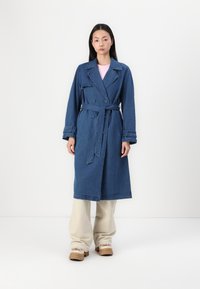 Denim plašč v temno modri barvi z dvojno gumbno zapenjanjem, širokim ovratnikom in pasom. Ima dolge rokave in ravno, dolgo krojenje.