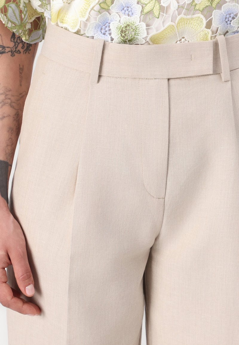 Beige maßgeschneiderte Hose aus einem strukturierten Stoff mit Falten an der Vorderseite und einem eleganten Bund. Hand ruht auf dem Oberschenkel.