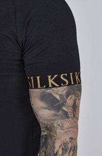 Schwarzes T-Shirt mit figurnaher Passform. Verfügt über ein goldfarbenes Logo-Band mit der Aufschrift "SILKSIK" auf dem Ärmel. Sichtbare Haut-Tattoos am Arm.