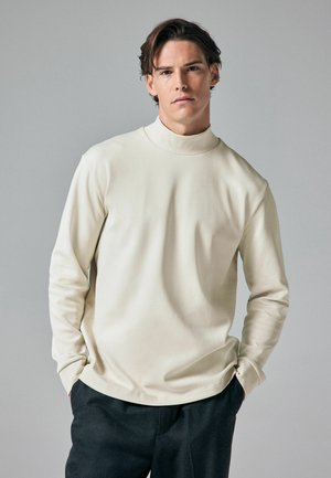 Bluză crem cu mâneci lungi și guler înalt, confecționată dintr-un material neted. Are o croială lejeră și un design minimalist.