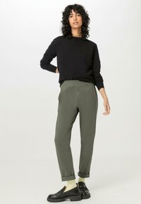 Schwarzer Pullover mit langen Ärmeln, kombiniert mit olivgrünen, schmal zulaufenden Hosen, die am Knöchel einen Saum haben, und getragen mit schwarzen Schuhen und hellen Socken.