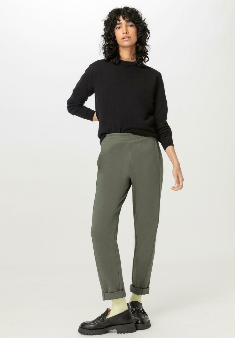 Schwarzer Pullover mit langen Ärmeln, kombiniert mit olivgrünen, schmal zulaufenden Hosen, die am Knöchel einen Saum haben, und getragen mit schwarzen Schuhen und hellen Socken.