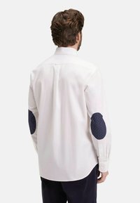 Chemise blanche à manches longues avec un col, ornée de coudières à pois bleu marine et de poignets boutonnés. Texture lisse, coupe ajustée.