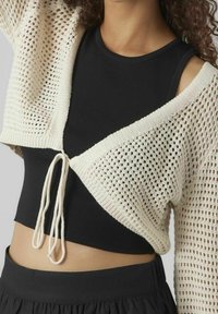 Žena v béžovém krátkém pleteném kardiganu zavázaném vpředu přes černý bezrukávový crop top a černou sukni.