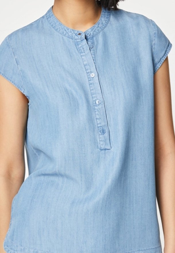 Femme portant une chemise sans manches en chambray bleu clair avec un col mandarin et une patte de boutonnage à quatre boutons sur le devant.