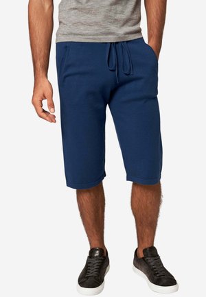 Mann trägt marineblaue, mit Kordelzug versehene, knielange Shorts, graues Hemd und schwarze Sneaker mit weißen Sohlen, steht mit entspannten Händen.