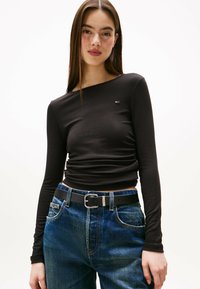 Schwarzes Langarmshirt mit Rundhalsausschnitt und dezentem Logo-Detail. Kombiniert mit blauen Denim-Jeans und einem schwarzen Gürtel mit metallischer Schnalle.
