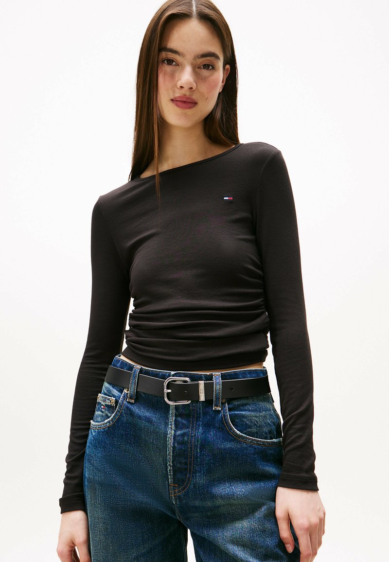 Schwarzes Langarmshirt mit Rundhalsausschnitt und dezentem Logo-Detail. Kombiniert mit blauen Denim-Jeans und einem schwarzen Gürtel mit metallischer Schnalle.