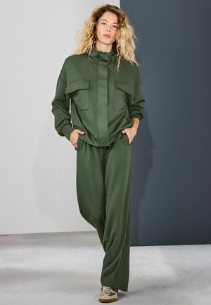 Ung kvinna med lockigt blondt hår klädd i en olivgrön träningsdräkt med fickor, stående med händerna i fickorna mot en tvåtonig grå bakgrund.