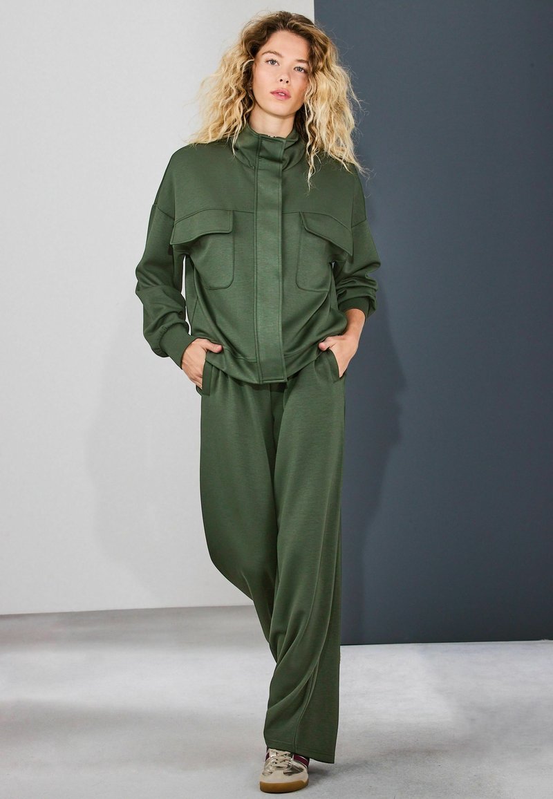 Young woman with curly blonde hair wearing an olive green tracksuit with pockets, standing with hands in pockets against a two-tone gray background.
