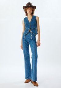Chaleco de denim con escote en V y cierre de botones, combinado con pantalones acampanados. Ambas piezas presentan costuras visibles y un color azul claro. Sombrero y botas marrones.