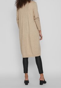 Cardigan lungo beige realizzato in tessuto morbido con orlo e polsini a coste. Abbinato a leggings neri, con un design elegante e affusolato.