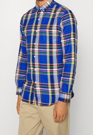 Homme portant une chemise à carreaux bleu multicolore à boutons avec un pantalon beige, debout les bras détendus le long du corps sur un fond neutre.
