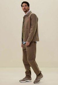 Bruine canvasjas met wolkenborduring, gelaagd over een groene shirt, gecombineerd met bijpassende bruine broek en grijze sneakers. Effen achtergrond.