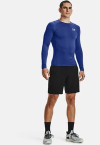 Langærmet blå træningsskjorte, sorte shorts og grå sneakers med sorte sokker. Skjorten har en glat tekstur og en tætsiddende pasform.