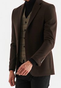 Blazer marrone su misura sopra un gilet a righe con bottoni. Il blazer ha una vestibilità slim e rever affilati, abbinato a un dolcevita nero.