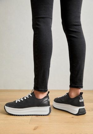 Osoba nosząca czarne obcisłe dżinsy i czarne sneakersy na platformie z białymi podeszwami stojąca na drewnianej podłodze.