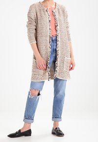 Stickad cardigan med ett strukturerat mönster i beige och krämfärgat, med en fransig framdel, långa ärmar och en ficka. Kombineras med slitna blåjeans.