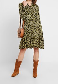 Svart klänning med gult blommönster, lager-på-lager design, långa ärmar, krage. Matchas med en brun lädercrossbody-väska och bruna stövlar.