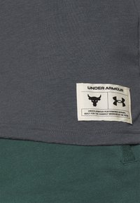 Camiseta de algodón gris con una etiqueta que presenta un logo de toro negro, marca Under Armour, y tejido verde en el borde inferior, que sugiere superposición.