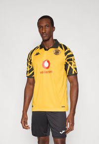 Κίτρινη αθλητική φανέλα με μαύρα μανίκια με σχέδια, που διαθέτει γιακά, λογότυπα χορηγών και σήμα συλλόγου. Συνδυάζεται με μαύρο σορτς.