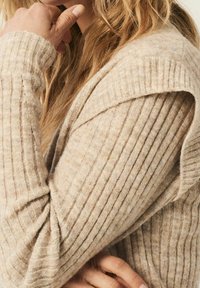 Pull beige en tricot avec texture côtelée, présentant un large col et une coupe décontractée. Matière douce et confortable avec des variations subtiles de couleur.