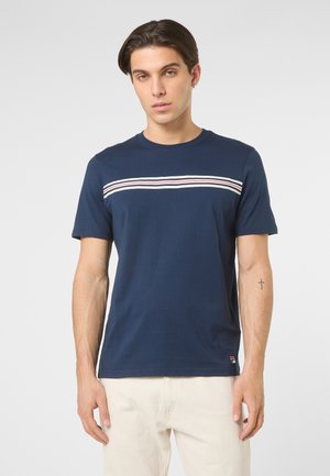 Junger Mann trägt ein marineblaues T-Shirt mit weißen und roten horizontalen Streifen über der Brust und hellbeige Hosen, steht vor einem schlichten Hintergrund.