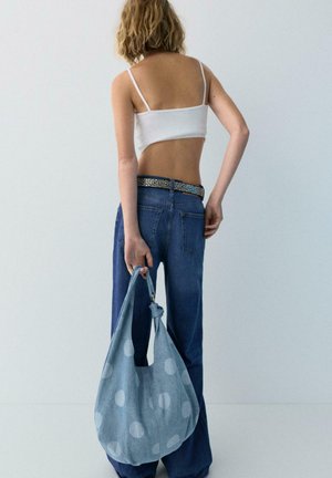 Femme en haut court blanc et jean bleu tenant un grand sac à bandoulière bleu à pois, debout devant un arrière-plan clair uni, de dos à la caméra.