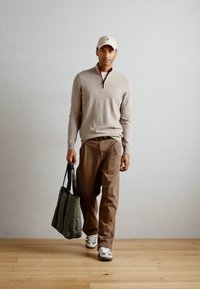 Beige zip-neck sweater, brown trousers, carrying a green quilted tote bag, and wearing white sneakers with green accents.