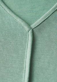 Světle zelený pletený top s výstřihem do V, který má jemnou texturu a kontrastní švy podél výstřihu a švů. Materiál je měkký a elastický.