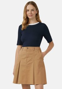 Vrouw met lichtbruin haar draagt een donkerblauwe top met korte mouwen en een hoge taille beige geplooide rok, staand met één hand in de zak tegen een grijze achtergrond.