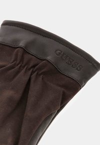 Bruine stoffen handschoen met een zwarte leren manchet. Heeft een geplooid ontwerp bij de pols en een gegraveerd "GUESS"-logo op de manchet.