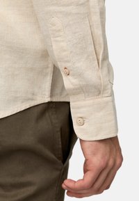 Chemise beige à manches longues avec un motif rayé subtil, présentant des poignets boutonnés et des ourlets arrondis. Porte avec un pantalon vert.