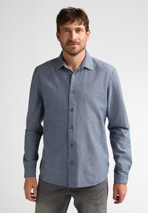 Mann mit kurzen braunen Haaren und Bart, der ein blaues, langärmeliges Hemd mit Knöpfen und graue Hosen trägt, steht vor einem schlichten Hintergrund.