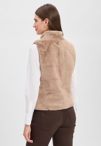 Beżowa kamizelka z futra syntetycznego z wysokim kołnierzem, połączona z białą koszulą z długim rękawem. Tył ma fakturę, bez widocznych zapięć.