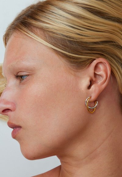 Close-up zijaanzicht van een persoon met blond haar die verweven gouden en zilveren oorringen draagt aan het linker oor.