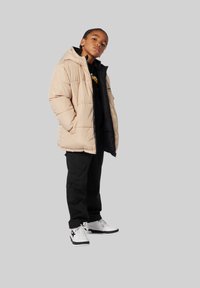 Doudoune beige avec capuche, couche intérieure noire, pantalon cargo noir et baskets blanches avec accents noirs. Posture droite, mains dans les poches.