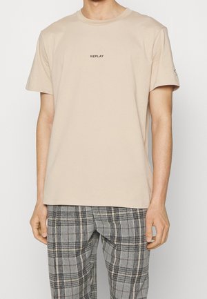 Beige cotton crew-neck t-shirt featuring a small black "REPLAY" logo on the chest, paired with gray plaid trousers.