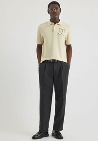 Beige Poloshirt mit schwarzem "N"-Logo, kombiniert mit grauen Hosen und schwarzen Schuhen. Glattes Material, klassischer Kragen und taillierte Passform.