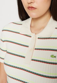 Femme portant un polo à manches courtes côtelé avec une base crème et des rayures horizontales vertes, rouges et jaunes, arborant un petit logo de crocodile vert.