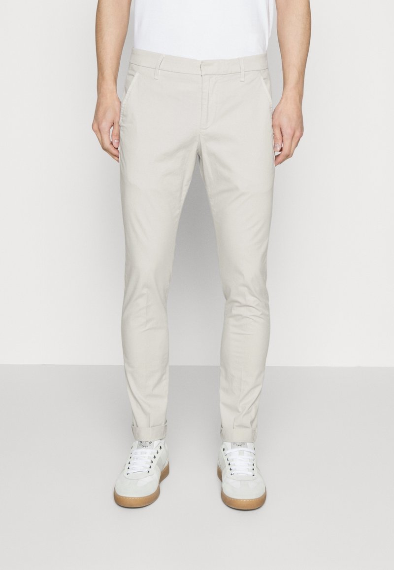 Homme portant un pantalon beige ajusté avec ourlets retroussés et baskets blanches, debout devant un fond clair uni.