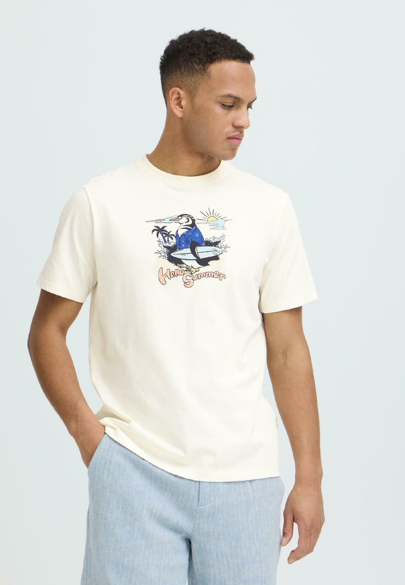 Homme portant un t-shirt crème avec un motif de surf d'oiseau et le texte "Hello Summer", associé à un pantalon bleu clair, debout devant un fond uni.