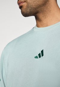 Gaiši zila kokvilnas T-krekls ar tumši zaļu logotipu kreisajā krūšu pusē. Audums ir gluds, ar ikdienišķu apaļu kakla dizainu.