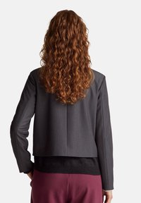 Giacca grigia su misura con design corto, tessuto texturizzato, revers a punta e maniche lunghe, abbinata a un maglione nero e pantaloni bordeaux.